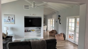 Una sala de estar con televisor de pantalla plana, un sofá, una mesa de centro de vidrio y un ventilador de techo.