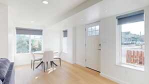 Une pièce moderne et bien éclairée, avec une table et des chaises, de grandes fenêtres donnant sur une rue et sur de la verdure, et une porte menant à une autre pièce.