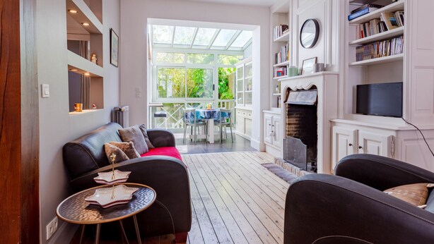 Un salon moderne avec un canapé, une petite table ronde et une cheminée. On y trouve une bibliothèque remplie de livres et un téléviseur. Une grande fenêtre laisse entrer la lumière naturelle et offre une vue sur la verdure à l’extérieur.