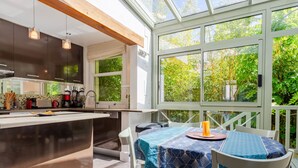 Une cuisine moderne dotée d’un grand îlot central, d’un coin repas avec une table et des chaises, et offrant une vue sur la verdure grâce à de grandes fenêtres.