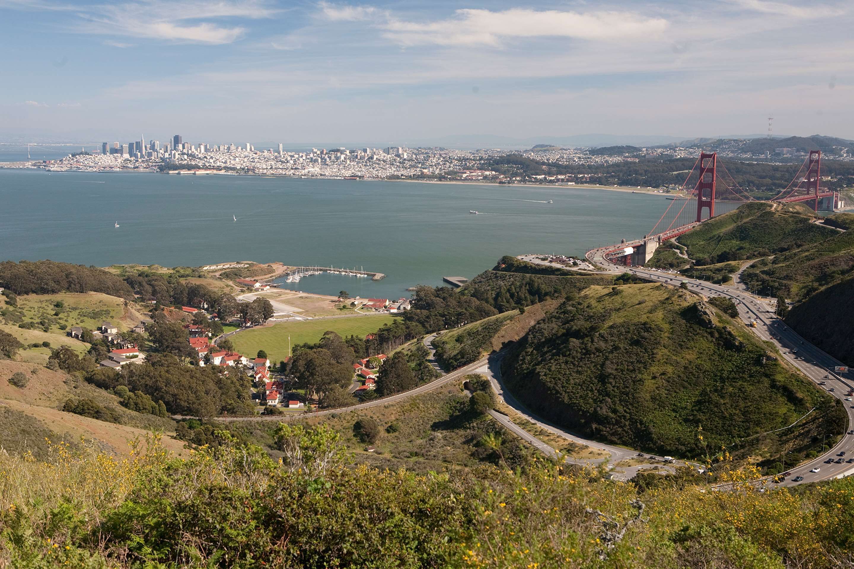 Un paisaje urbano costero con un puente rojo destacado, colinas verdes y un puerto deportivo.