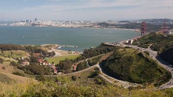 Un paisaje urbano costero con un puente rojo destacado, colinas verdes y un puerto deportivo.