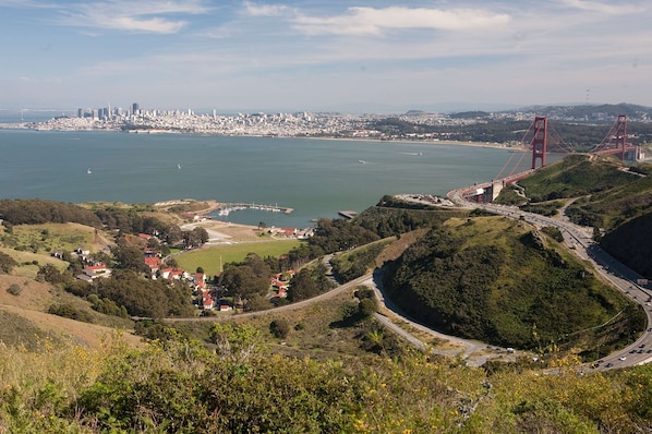 Un paisaje urbano costero con un puente rojo destacado, colinas verdes y un puerto deportivo.