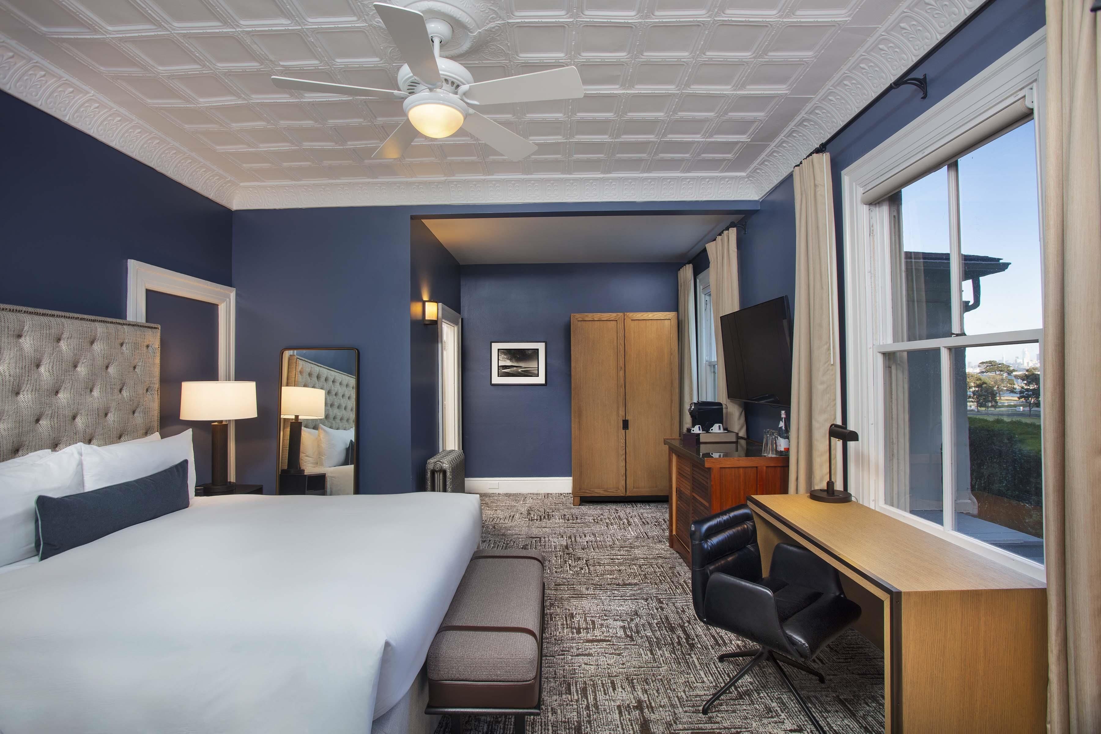 Habitación de hotel con cama, escritorio, silla, ventilador de techo y ventana grande.