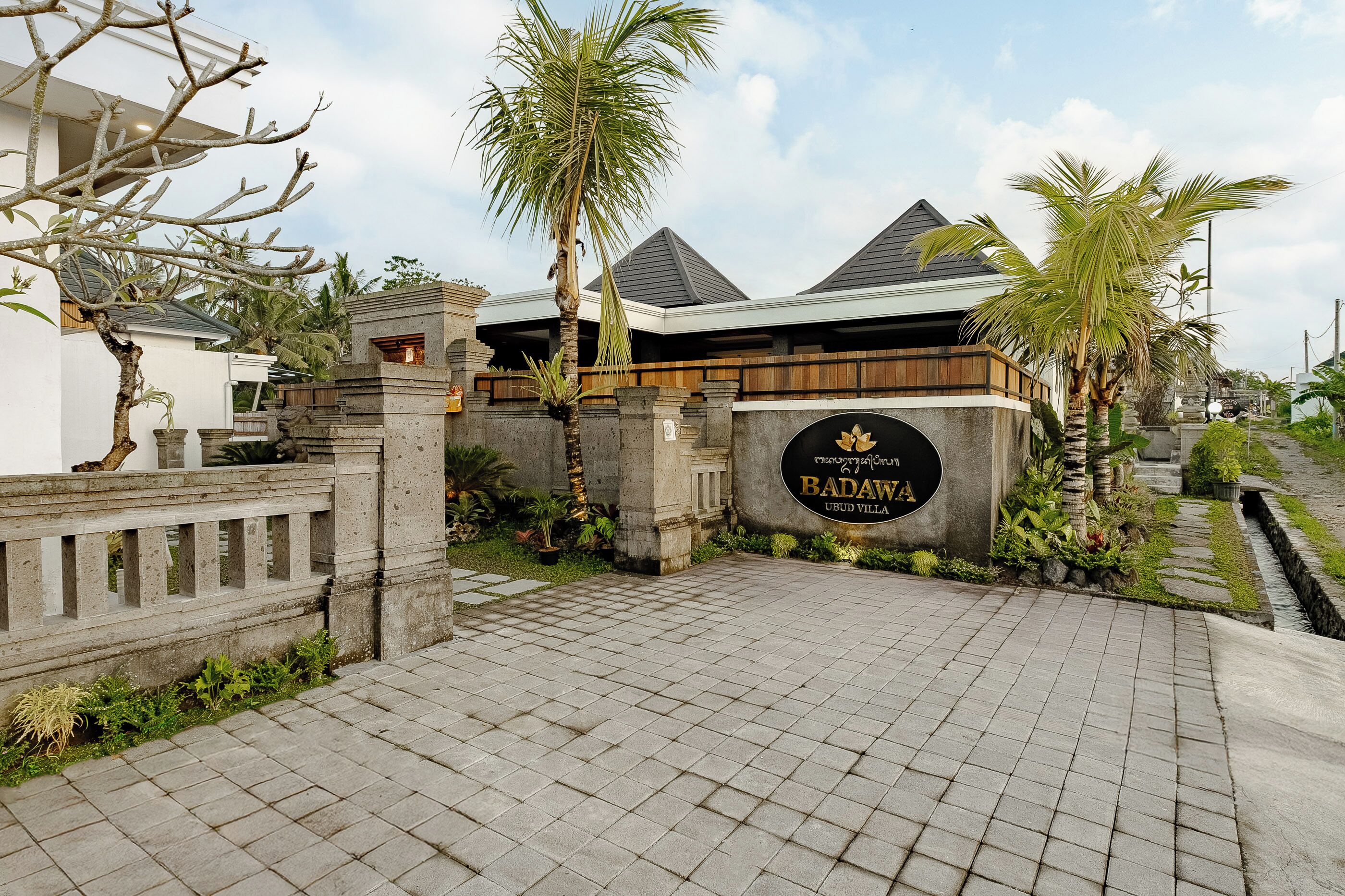 A paved entrance to Badawah Ubud Villa with a stone wall, palm trees, and a signboard.