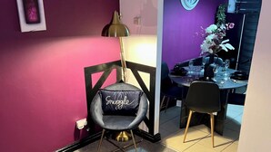 A room with a purple wall, a black chair with a cushion labeled "Snuggle," a table with a vase of flowers, and a clock on the wall. - 2-Bed - Sleeps 4 - Pet Friendly & Free Parking (Bolton)