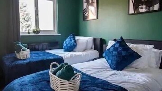 A room with two beds, each with a blue bedspread and white pillows, a window with a view of trees, and a wall with two framed pictures. - 2-Bed - Sleeps 4 - Pet Friendly & Free Parking (Bolton)