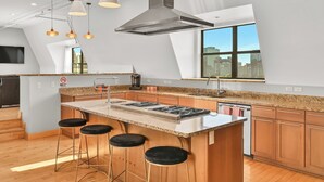 A modern kitchen with a central island, bar stools, a stainless steel range hood, pendant lights, and a large window offering a city view. - Penthouse • Fiserv, Bars, Perks & Big City Views • Downtown Milwaukee (Milwaukee)