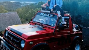 Two people sitting in a red Suzuki car with a view of a mountain and a river in the background. - Kintamani Angel (Kintamani)
