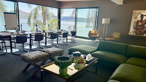 A spacious living area with a large window overlooking a lake, a dining table with chairs, a green sofa, and a coffee table with fruit and a decorative bowl. - Peats Bite  (Berowra Waters)