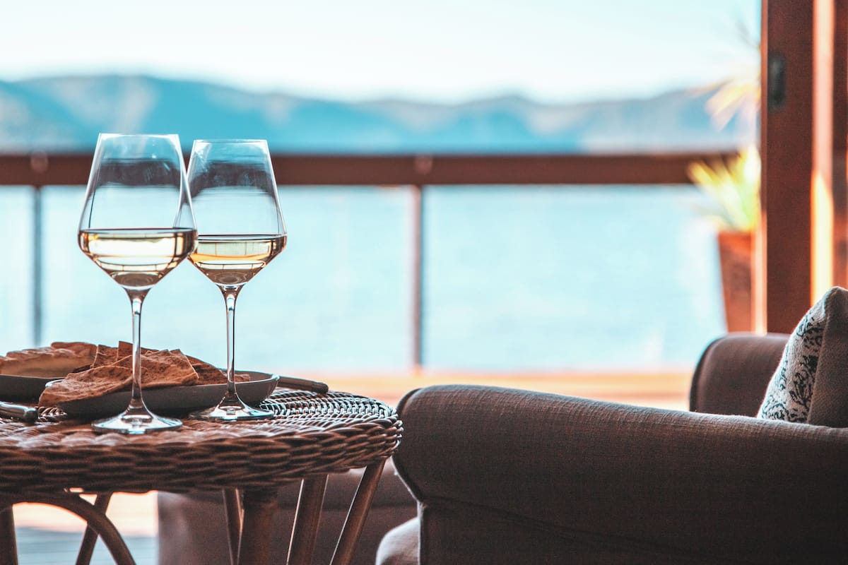 Two wine glasses on a wicker table with a view of the sea and mountains in the background.