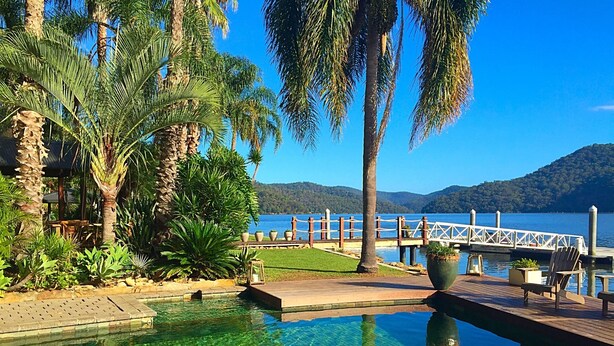 A clear pool with a wooden deck, surrounded by palm trees and a view of a lake and mountains. - Peats Bite  (Berowra Waters)