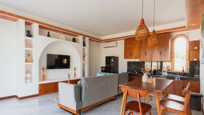 A modern kitchen with wooden cabinets, a dining area with a table and chairs, and a grey sofa. - Karang Padma Villas (Kutuh)