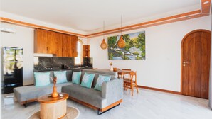 A modern living room with a grey sectional sofa, wooden dining table, and a kitchen area with wooden cabinets and a painting on the wall. - Karang Padma Villas (Kutuh)