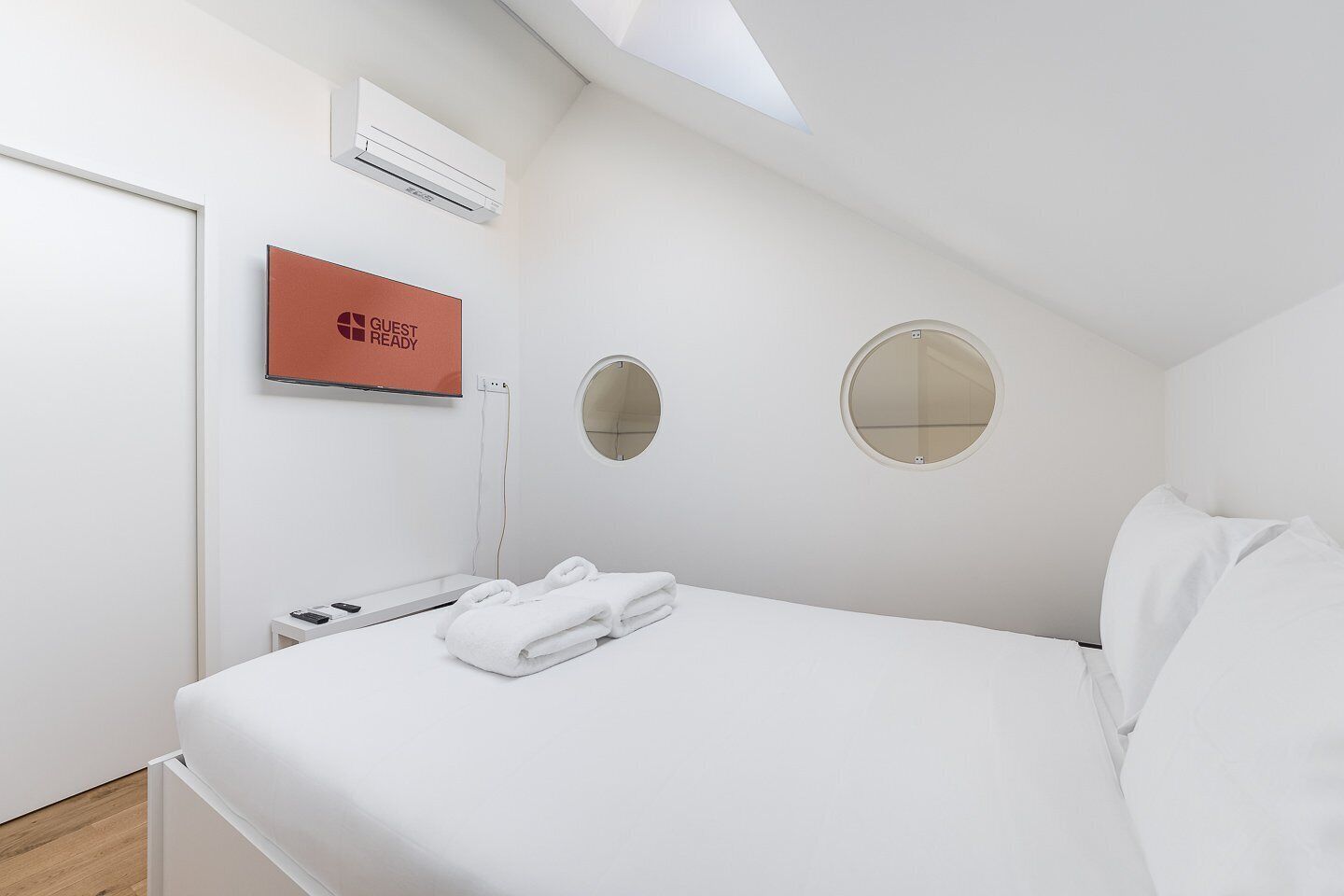 Una camera d'albergo con un letto, un condizionatore d'aria a muro, un cartello con scritto 'GUEST READY', due specchi rotondi e un asciugamano bianco piegato ordinatamente sul letto.