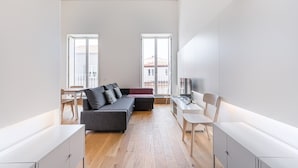 Een moderne, minimalistische woonkamer met een grijze bank, houten vloeren en een televisie op een witte kast.