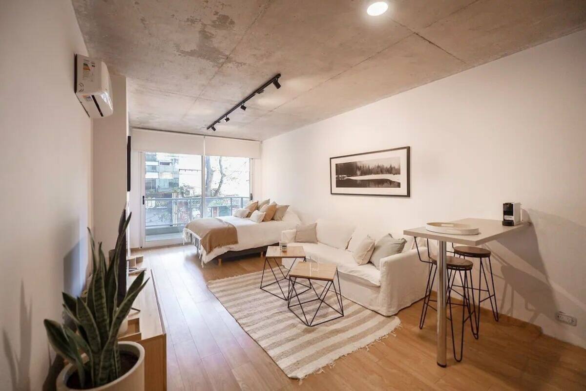 A modern, minimalist living room with a white sofa, a dining area with stools, a large window, and a potted plant.