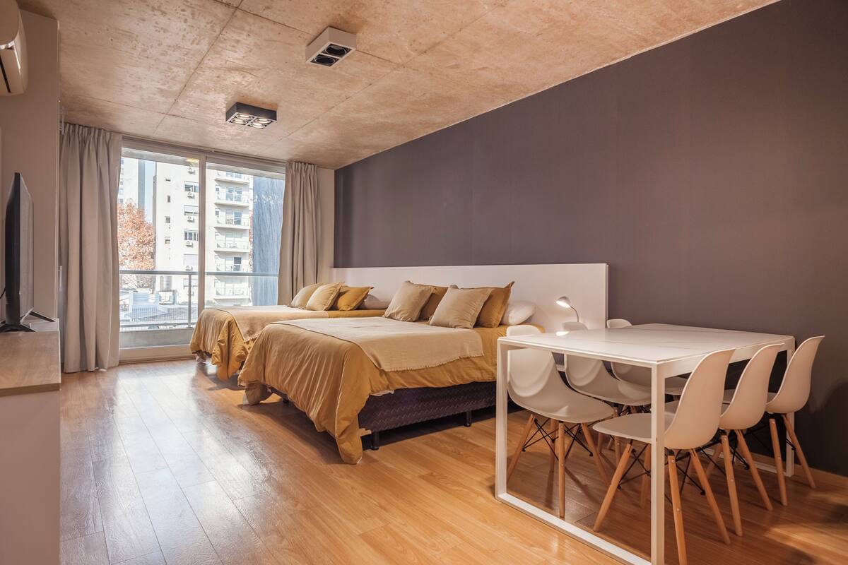 A modern bedroom with a large bed, a dining table with chairs, and a view of the city through a large window.