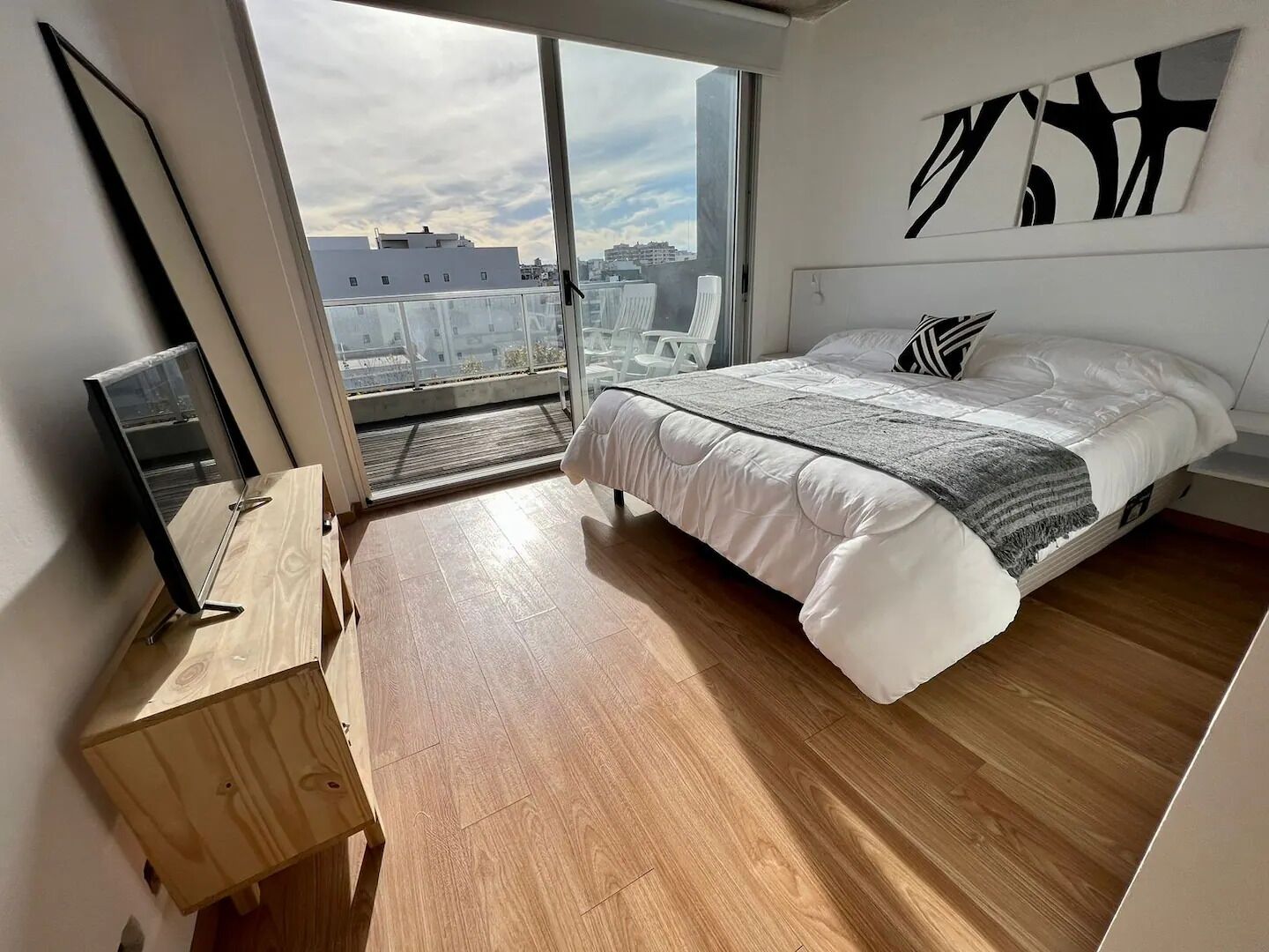 A modern bedroom with a large bed, a wooden floor, a city view through a sliding door, and a mounted artwork on the wall.