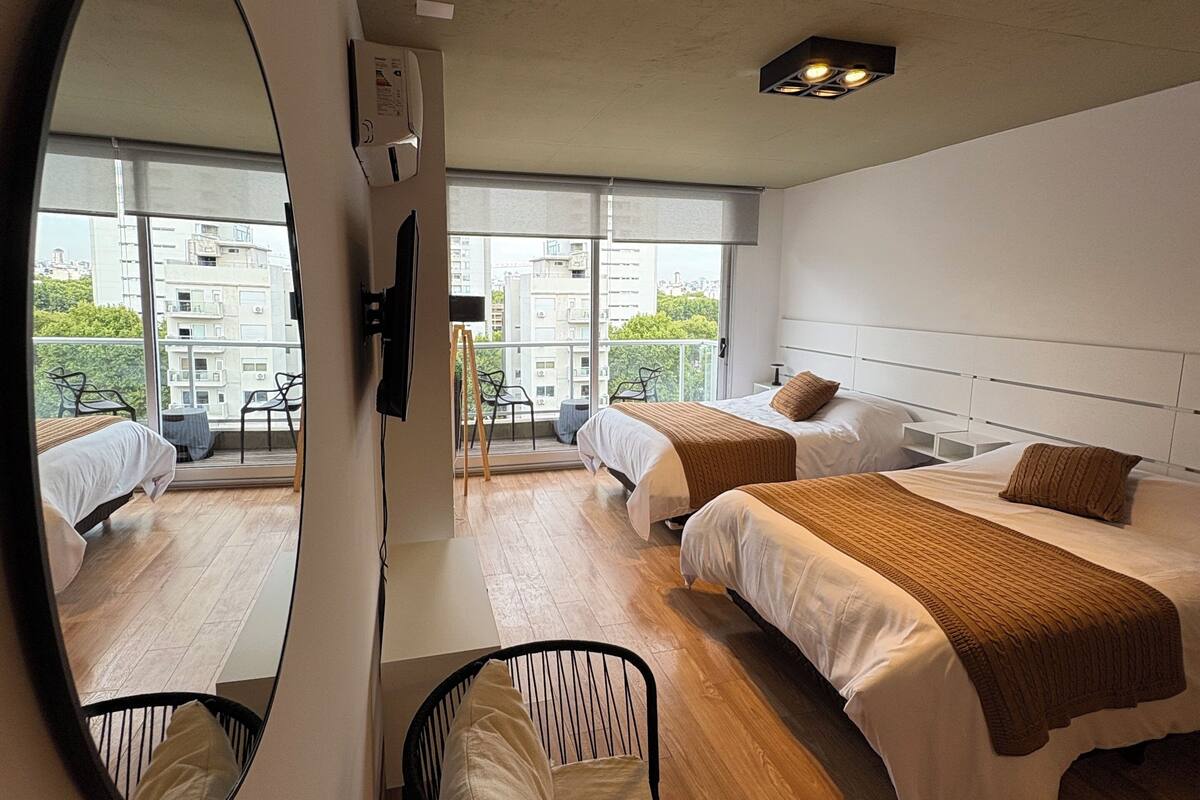 A modern hotel room with two beds, a large mirror, a small table, and a view of the city through large windows.