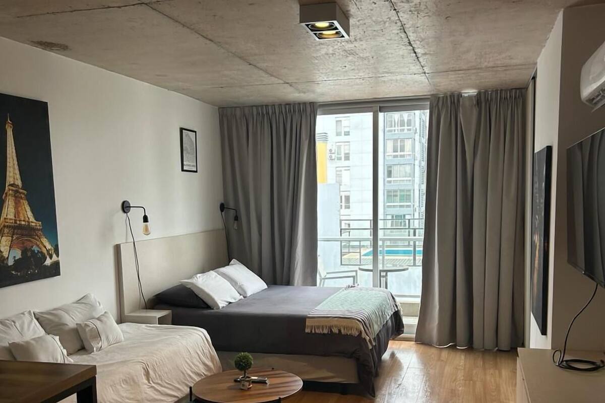 A modern hotel room with a large bed, a small table, a TV, and a view of the city through large windows.