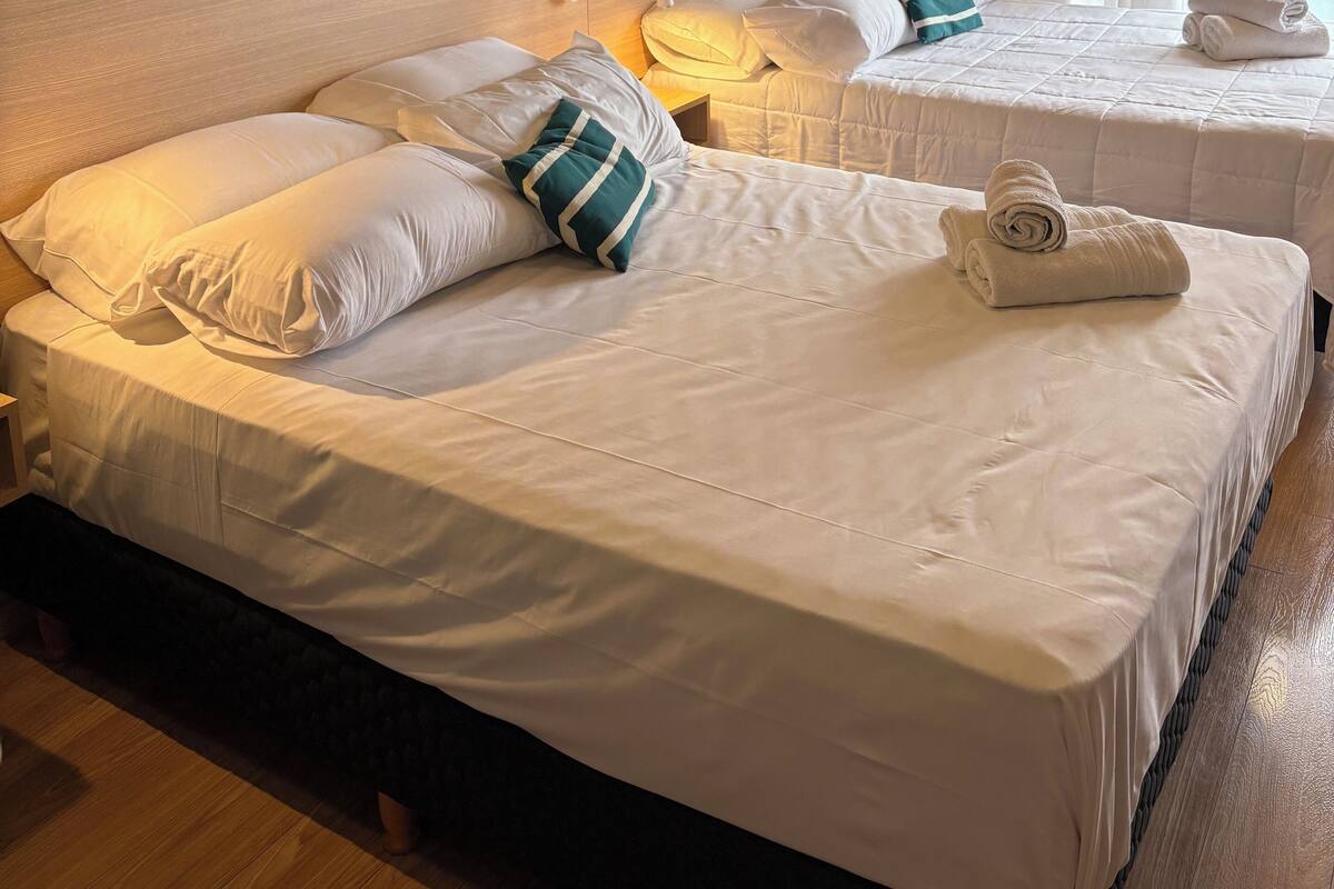 A hotel room with two beds, a headboard, wall art, a window with curtains, and a neatly made bed with a towel.