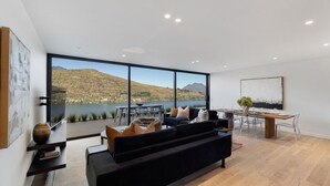 A modern living room with a large window overlooking a lake and mountains. There is a black sofa, a dining table with chairs, and a large painting on the wall. - The Bunker Charlie A-2 Bedroom (Queenstown)