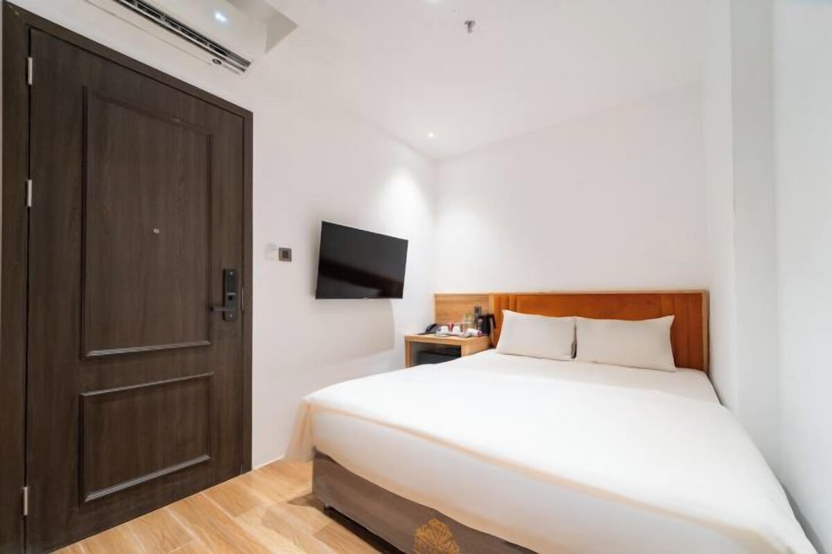 A hotel room with a large wooden door, a flat-screen TV mounted on the wall, a wooden headboard, and a neatly made bed with white linens.