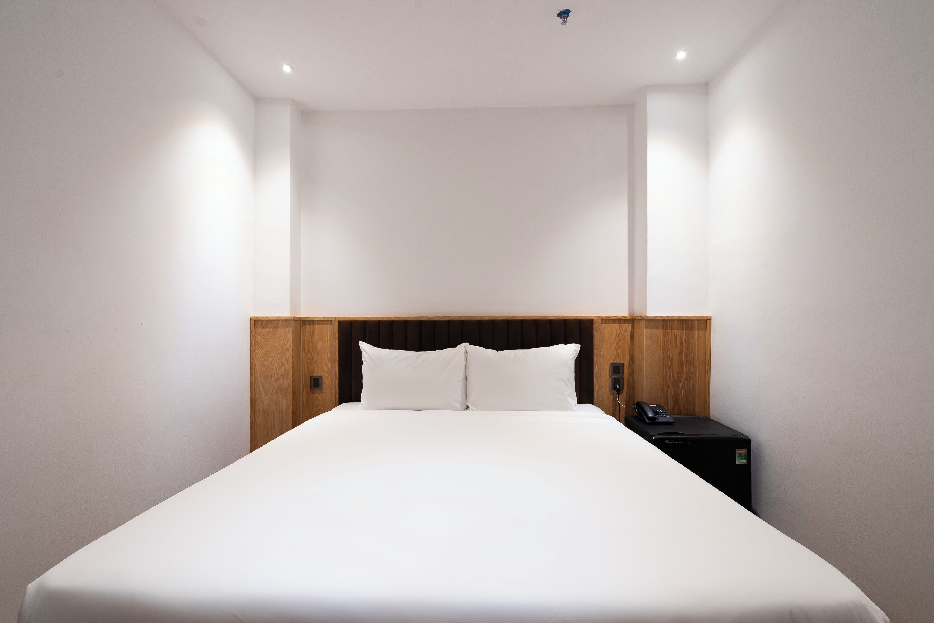 Un lit bien fait dans une chambre d’hôtel, avec une tête de lit en bois, une table de chevet sur laquelle se trouve un téléphone et un petit appareil noir.