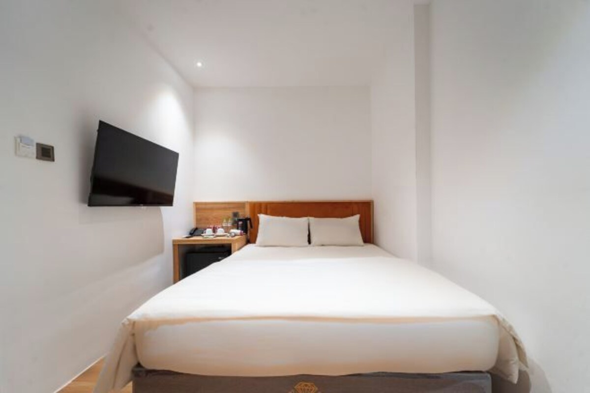 A hotel room with a single bed, a flat-screen TV mounted on the wall, a bedside table with a lamp, and a wooden headboard.