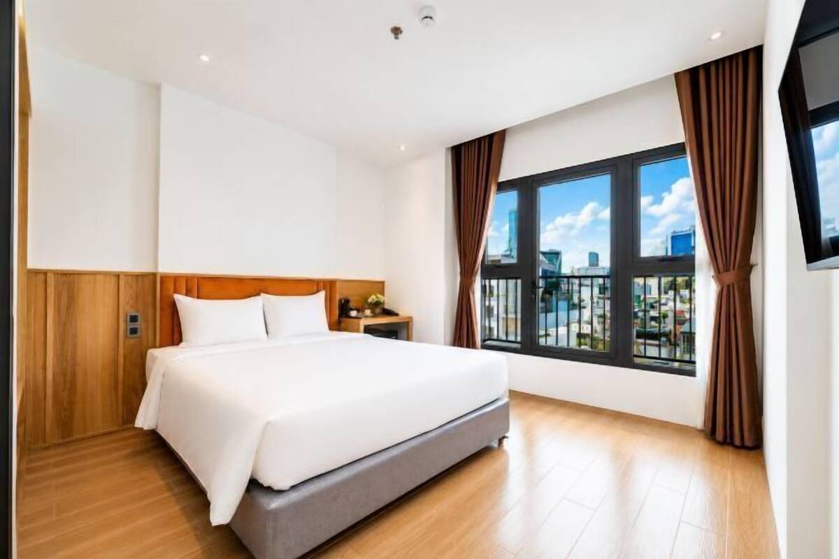 Una habitación de hotel con una cama grande, paneles de madera, una mesita de noche, un televisor y una ventana con cortinas.