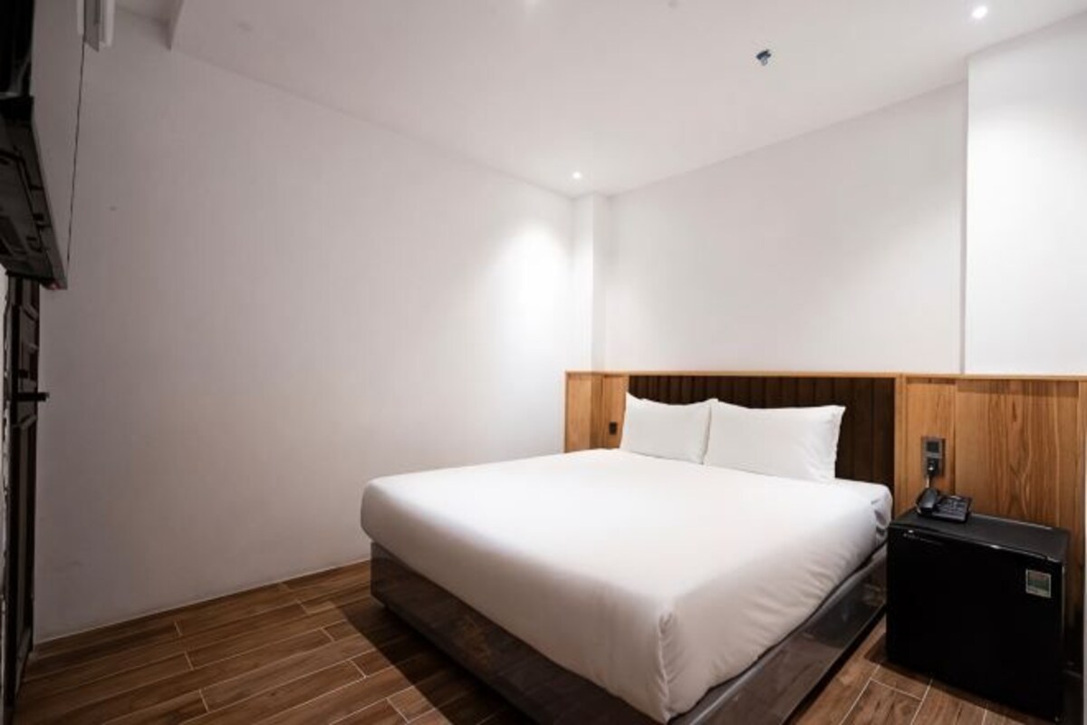 A hotel room with a large bed, wooden headboard, a bedside table with a lamp, and a small black nightstand.
