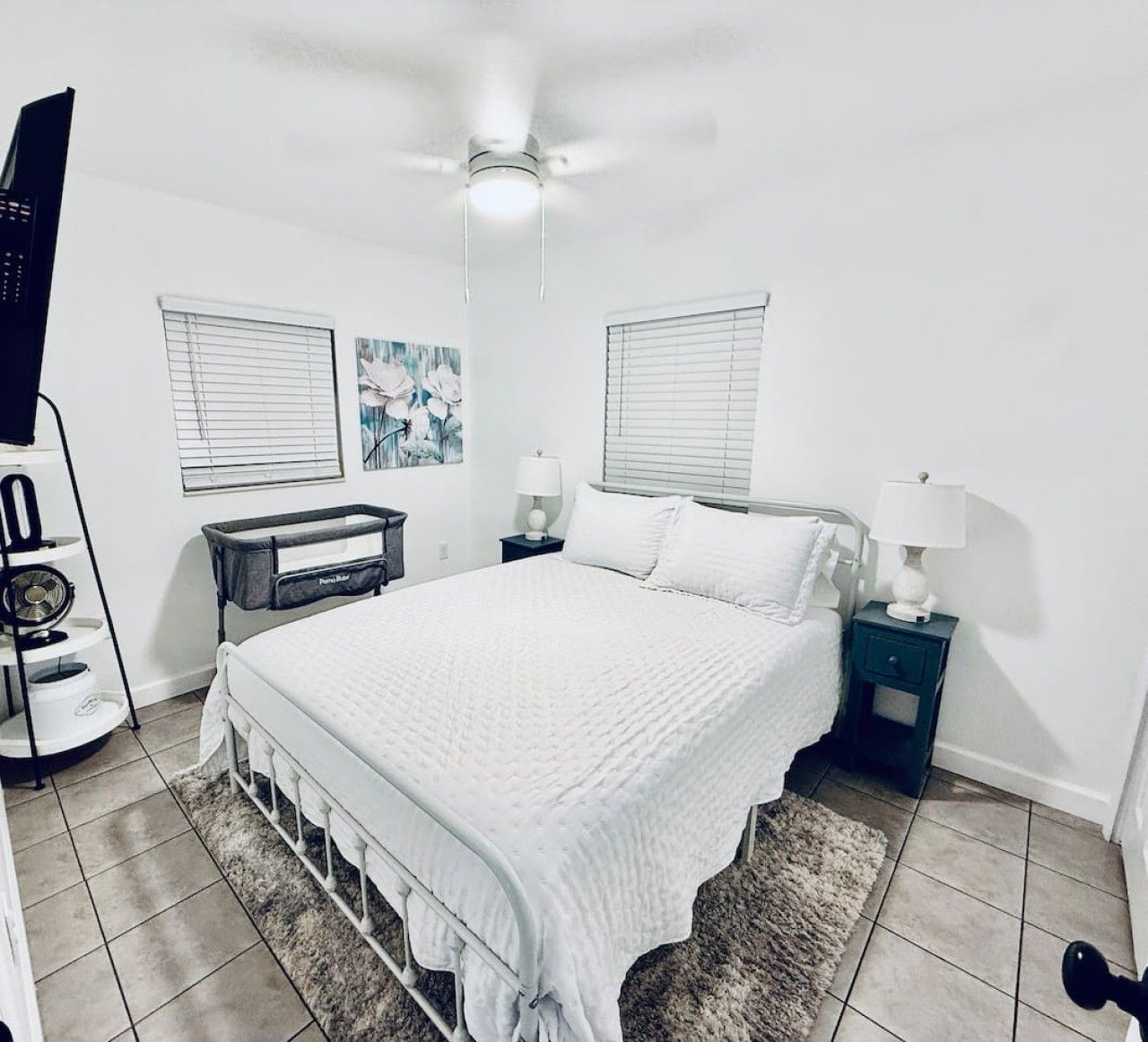 Une chambre à coucher comprenant un lit, deux tables de chevet, un ventilateur de plafond et une fenêtre avec des stores.