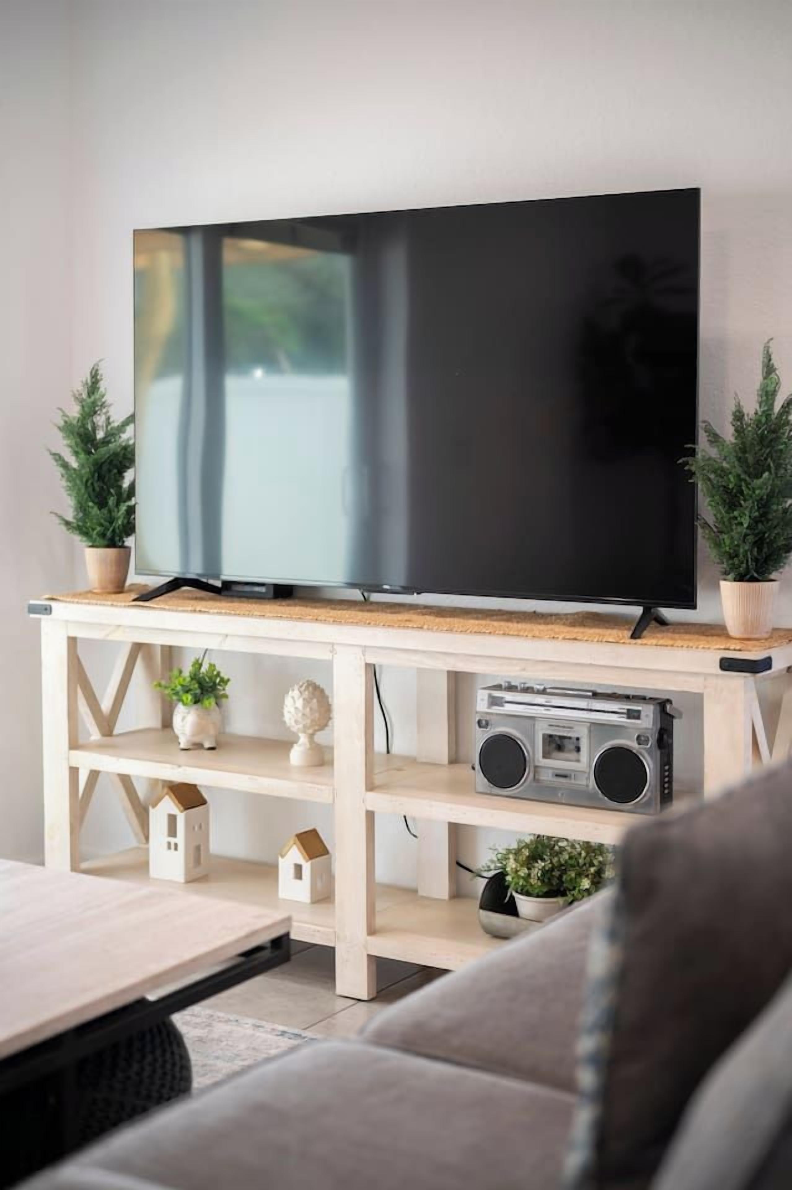 Litteänäyttöinen televisio, joka on sijoitettu puiseen TV-telineeseen, jossa on hyllyjä, sekä kannettava stereolaite ja pieniä, taloa muistuttavia koriste-esineitä.