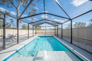 A covered swimming pool with a clear blue water feature, surrounded by a metal frame structure, and a view of the backyard with a fence and trees.