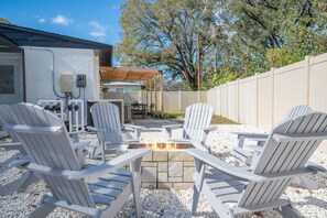 Un espace patio aménagé avec des fauteuils Adirondack disposés autour d’un foyer, un mur en briques blanches et une pergola en bois.