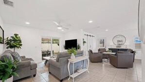Een moderne woonkamer met een grijze bank, een salontafel en een televisie. Er staan planten, er is een plafondventilator en een eethoek is zichtbaar.