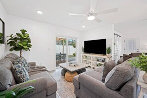 Un salon moderne avec des canapés gris, un téléviseur à écran plat, un ventilateur de plafond et un coin repas comprenant une table et des chaises.