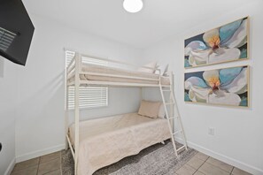 Une chambre avec un lit superposé, une échelle, une fenêtre avec des stores et deux tableaux aux motifs floraux accrochés au mur.