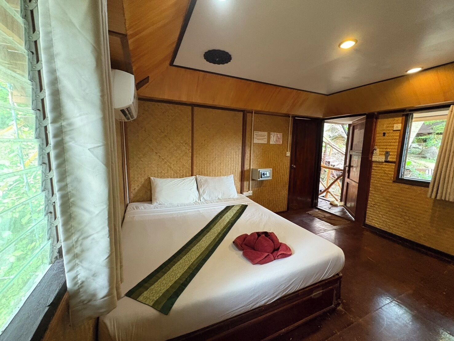 Habitación de hotel con cama, ventana con cortinas, puerta que da a un espacio exterior y un equipo de aire acondicionado instalado en la pared.