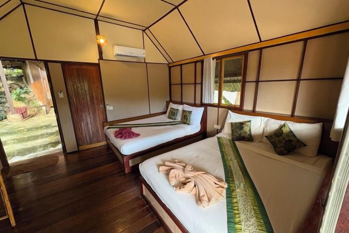 A room with two beds, wooden flooring, a window with curtains, and an air conditioning unit.