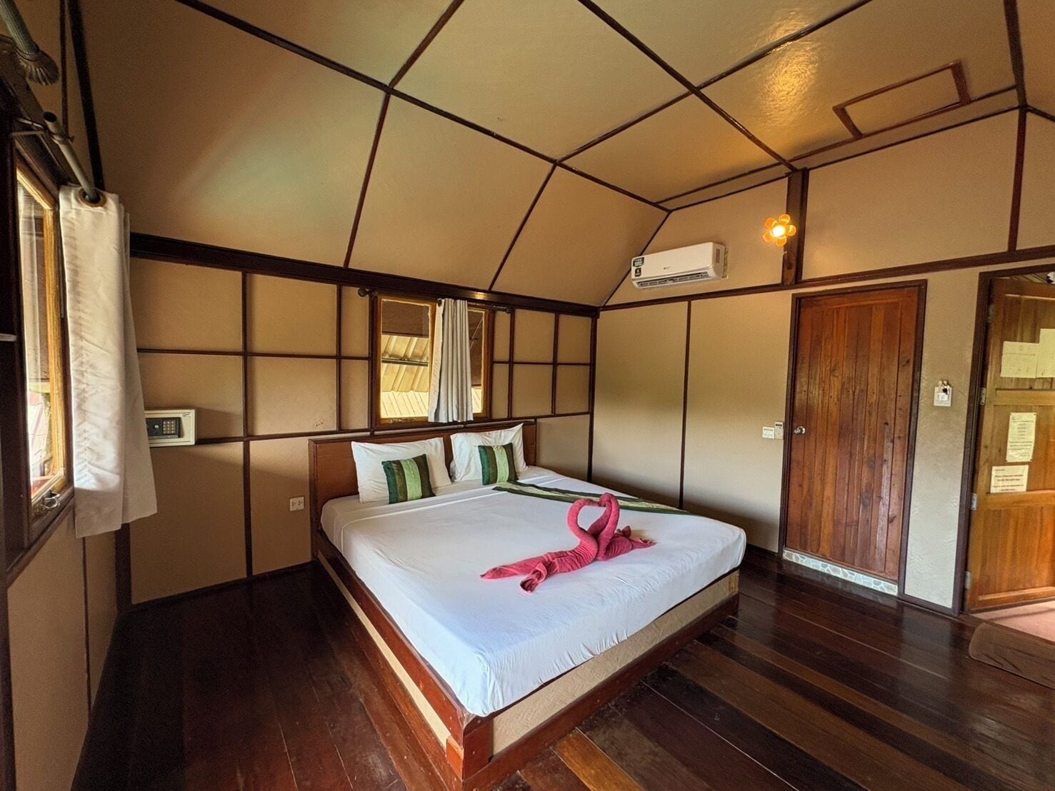 Quarto de hotel com uma cama grande, piso de madeira, janela com cortinas, ar condicionado e porta de madeira.
