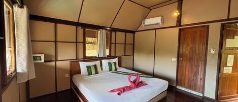 A hotel room with a large bed, a wooden floor, a window with curtains, an air conditioning unit, and a wooden door.