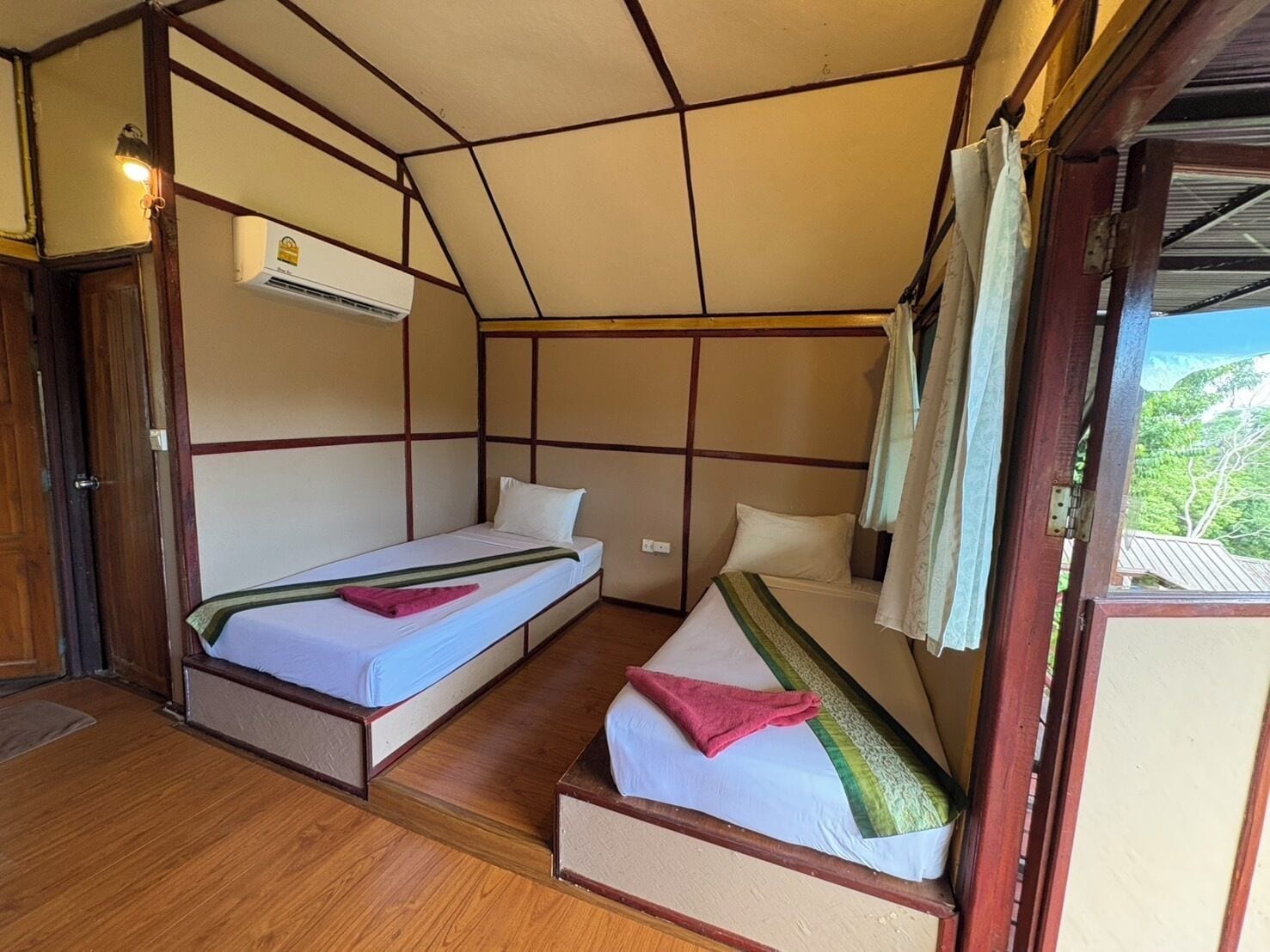A room with two beds, wooden flooring, an air conditioning unit, and a view of greenery outside.