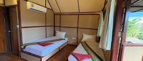A room with two beds, wooden flooring, an air conditioning unit, and a view of greenery outside.