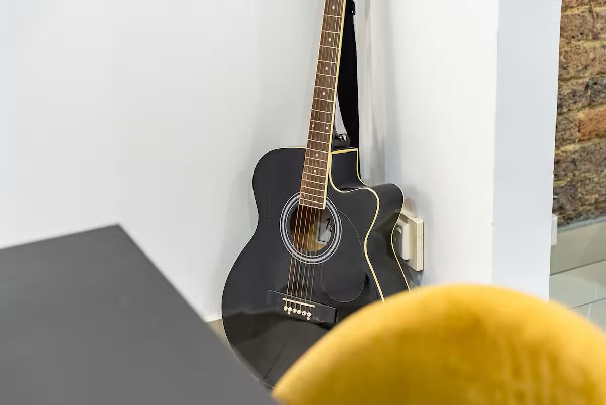 A black guitar leaning against a white wall in a room with a black table and a yellow chair.