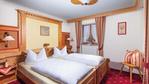 Una cama doble con sábanas blancas, un cabecero de madera, una mesita de noche con una lámpara, una pequeña mesa con una botella y una ventana con cortinas.