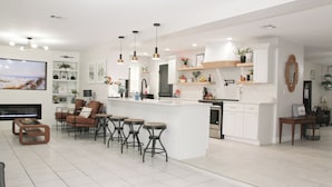 A modern kitchen with white cabinetry, a central island with bar stools, pendant lighting, and a large flat-screen TV. - 3BR Open-Plan Retreat w/ Heated Pool, Screened Patio & Grill – Melbourne (Melbourne)