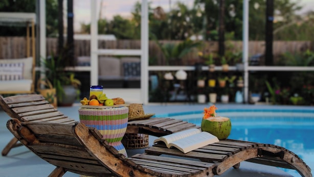 A poolside area with wooden lounge chairs, a wicker basket with fruits, and a book. - 3BR Open-Plan Retreat w/ Heated Pool, Screened Patio & Grill – Melbourne (Melbourne)