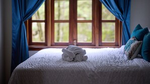 Una cama bien hecha con una toalla enrollada en el centro, una ventana con cortinas azules y una almohada con estampado azul y blanco.